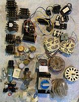 Photo shows a variety of vintage electrical components laid out on light fabric. Includes transformers, capacitors, plugs, analog meter, switches, and wiring.