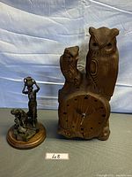 Photo showing hand-carved wooden owl clock alongside bronze figurine statue on wooden base on a grey surface with white backdrop