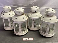 Four white Ikea candle lanterns with star cutouts on top and glass paneled sides with doors and rope handles lined up together