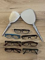 Top-down photo showing seven pairs of reading glasses arranged in two rows below a handheld mirror and brush set on a wooden surface.