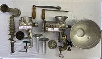 Photo showing set of two manual meat cutting machines and various metal accessories arranged on white tablecloth: spiral grinder, funnels, mixing bowl, grinder plates.