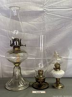 Three vintage oil lamps shown upright with glass chimneys and metal bases on a white background