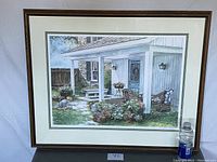 Framed lithograph showing the front porch of a home with potted plants, outdoor bench, rocking chair, and a child crouching on the grass.