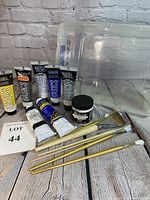 Full lot comprising 8 acrylic paint tubes, 7 brushes, and clear plastic storage bin on a grey surface against a brick backdrop