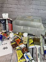Overview of acrylic paint tubes, brushes, sponges, and clear plastic tote on wooden surface against brick background