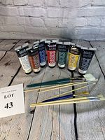 Seven tubes of Liquitex Basics acrylic paint arranged in a semicircle with seven paintbrushes laid in front on a wooden surface, white brick background.