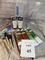 Complete view of acrylic paint tubes, brushes, markers, and plastic storage bin arranged on a white cloth background with brick wall.