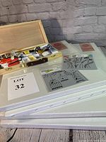 Overview photo showing wooden box of oil paints open with tubes visible, stacked canvases, and two stencils laid flat on top.