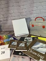 Photo showing art supplies lot with two blank canvases, multiple Strathmore drawing pads, Canson pastel pad, plastic carrying case and bubble sticks arranged on a wooden floor with a brick wall background.