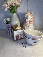 Full view of all items in the lot arranged with various vases, ceramic cookie jars, bowls, and baking molds together on a grey surface with a wood background.