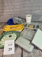 Photo showing the assortment of paint trays, mixing palettes, plastic storage containers, plates, and cups arranged on a surface with a brick patterned backdrop.