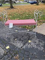 Full view of the white wrought iron bench outdoors on pavement with greenery and autumn leaves in background. Bench has a red and white checkered cushion seat and curved armrests.