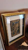 Framed floral wall art resting on a couch, showing gold decorative frame and layered three-dimensional floral relief inside.