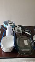 Wide-angle photo showing CorningWare casseroles with lids, square and round glass bakeware including Anchor Hocking, a white ceramic carafe, fry pans, and blue mixing bowls arranged on a table.