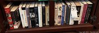 Books arranged on two shelves inside a wooden cabinet showing spines and titles, some with price tags