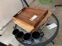 Photo showing wooden tray on top, two wooden cutting boards below, and three coffee mugs lined up in front on black round metal table.