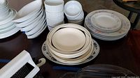 Multiple stacks of assorted plates, bowls, and ramekins in white and floral patterns displayed on a table in a garage.
