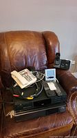 Photo showing Zenith DVD2200 disc player and Sony VCR stacked on a brown leather chair with multiple telephones, cables and a Seiko alarm clock on top.