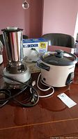 Photo of Hamilton Beach commercial bar mixer with stainless steel container next to Black & Decker Rice Cooker Plus on a wooden table.