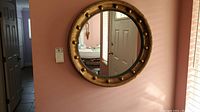 Round wall mirror mounted on a pinkish wall in hallway setting, reflecting a doorway and some furniture.
