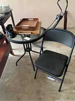 Glass top round metal outdoor table with black folding office chair nearby, wooden trays placed on table.