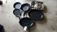 Photo showing seven pans on the floor, including roasting pans and frying pans with lids and grill patterns