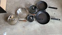 Top view of pot and pan set arranged on floor showing glass lids, wooden and black plastic handles, variety of sizes
