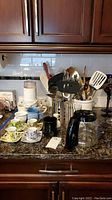 Kitchen counter with coffee pot, assorted cups and saucers, stainless steel utensil container with serving utensils, and a Salton coffee grinder visible.