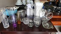 Photo shows assortment of clear and colored glass vases, pitchers, and a juicer on a table.