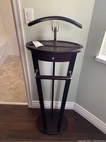Front view of dark wooden valet rack with curved hanger, small drawer, hanging bars, and round base.