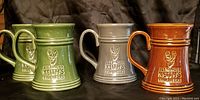 Four Alexander Keith's Fine Beer mugs in green, gray and brown colors, displayed side by side on a black fabric surface.