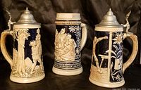 Front view of three beer steins showing detailed raised cream-colored relief on dark blue ceramic bodies, two lidded with pewter tops.