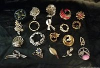 Photo showing 16 assorted brooches on a dark surface, various styles including floral, ribbon, leaf, and abstract shapes in gold and silver tones with stones and pearls.