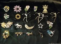 Photo showing all twenty assorted brooches arranged on a black surface, displaying various animal, floral, and decorative designs including angel, star, and dancing figures.