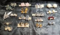 Full view of 15 different brooches and clip earrings arranged on black fabric.