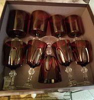 Nine cranberry coloured stemmed glasses laid out in box, showing four glasses of 4-4.5 inch height and five liqueur glasses approx. 5 inches tall with clear ornate stems.