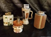 Four vintage decorative table lighters on black fabric background, showing different shapes and materials.