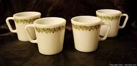 Four white Pyrex mugs each with green floral pattern around rim on dark background