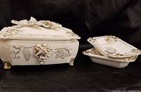 White ceramic trinket box with lid, decorated with gold grape cluster and leaf motifs, standing on gold-accented feet, shown with two matching small side plates.