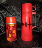 Two vintage thermoses standing side by side, one smaller with red and orange pattern, one larger red with black design and handle