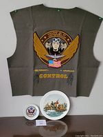 Full view of the embroidered jacket back patch pinned on a board with two porcelain commemorative plates placed below it