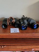 Two older binoculars side by side on wooden surface, showing front lenses and straps