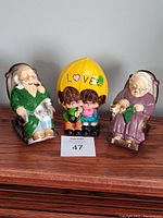 Front view of three ceramic coin banks: elderly man and woman on rocking chairs, and a couple under a yellow umbrella labeled 'LOVE'