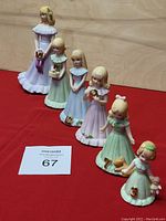 Six porcelain Enesco Growing Up Birthday Girls figurines arranged in a row on a red background, each holding a number or item corresponding to birthdays 3,7,9,10,11,16.