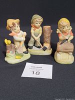 Front view of three porcelain figurines depicting children performing chores, against a black background with a lot number card.