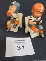 Photo of two porcelain football player figurines seated on white chairs, showing front and side angles with visible details.