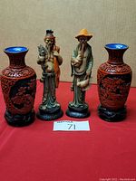 Front view of two Asian men statues placed between two cinnabar vases on a red cloth background.