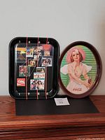 Two vintage Coca-Cola trays side by side, black rectangular tray with vintage advertising calendar on left, oval tray with illustrated woman on right.