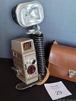 Bell & Howell 8mm movie camera with attached large flash light, brown carrying case, instruction manual and lot tag.