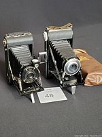 Two vintage folding cameras side by side, Kodak Kodo on left and Ansco on right, along with a leather pouch.